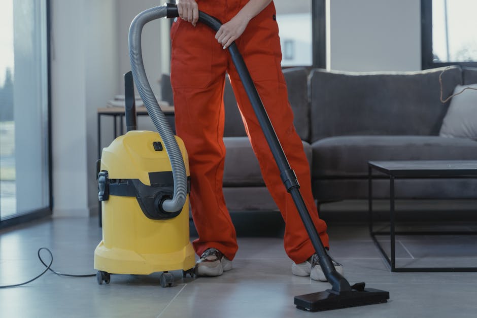 Close-up image showing a professional cleaning technician wearing a black glove using a handheld steam or deep cleaning machine to sanitise the grey carpeted floor inside a vehicle or residential spac